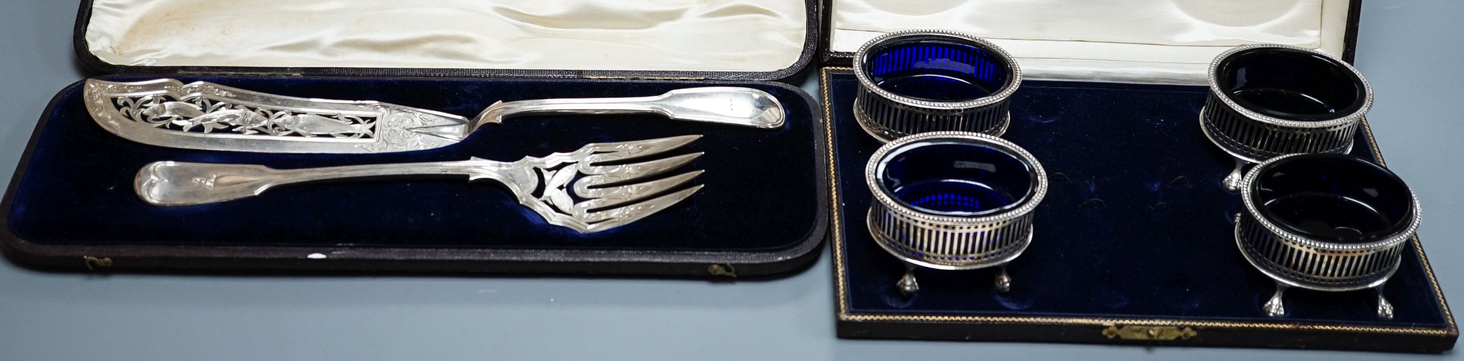A cased pair of Victorian silver fish servers, London, 1856, together with a cased set of four George V pierced silver oval salts with cobalt blue glass liners (no spoons).